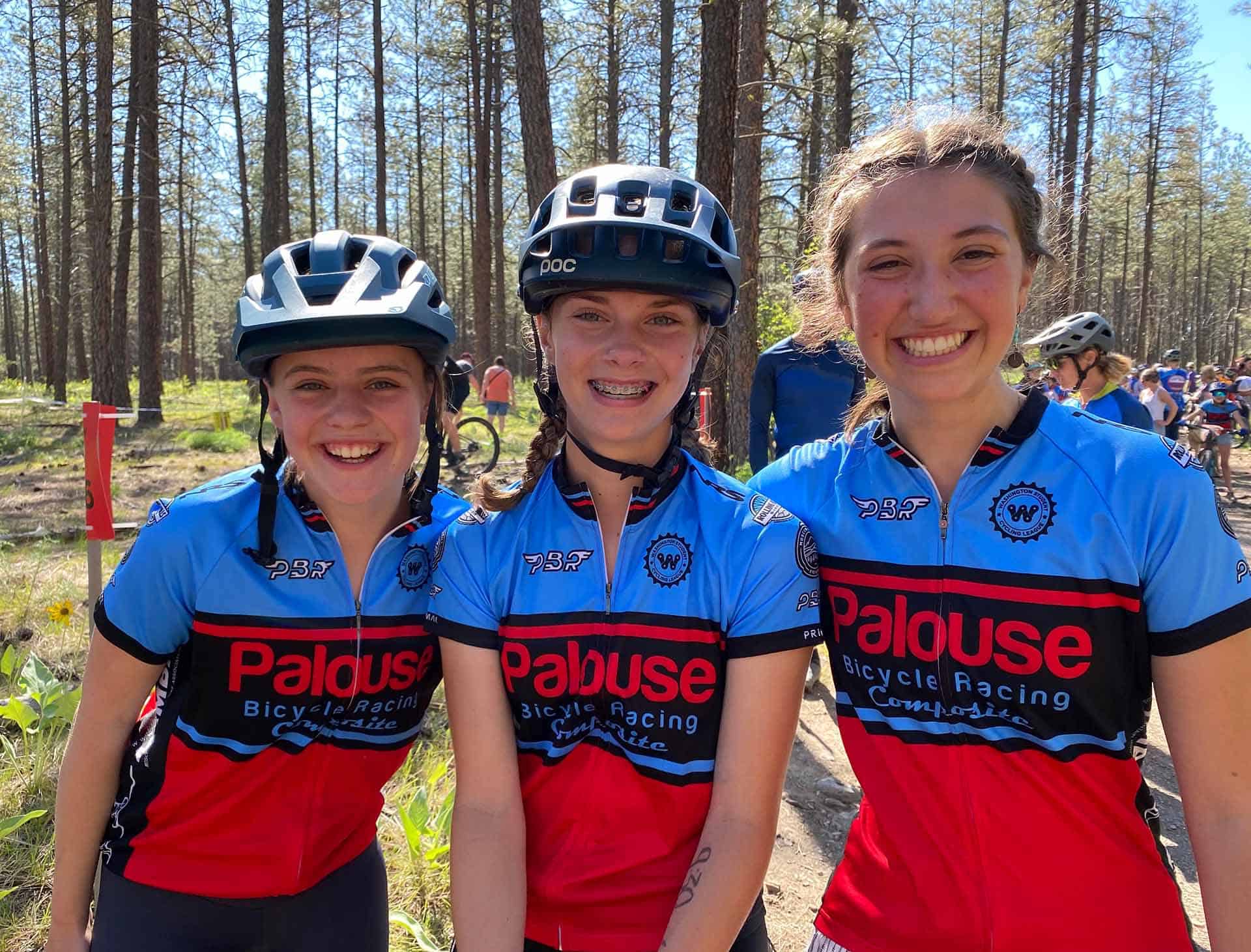 Three bicycle racers from the Palouse Bicycle Racing team smiling with forest race happening in the background