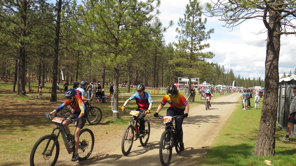 Four cyclists on trail at beginning of race with lots of people and tents in the distance