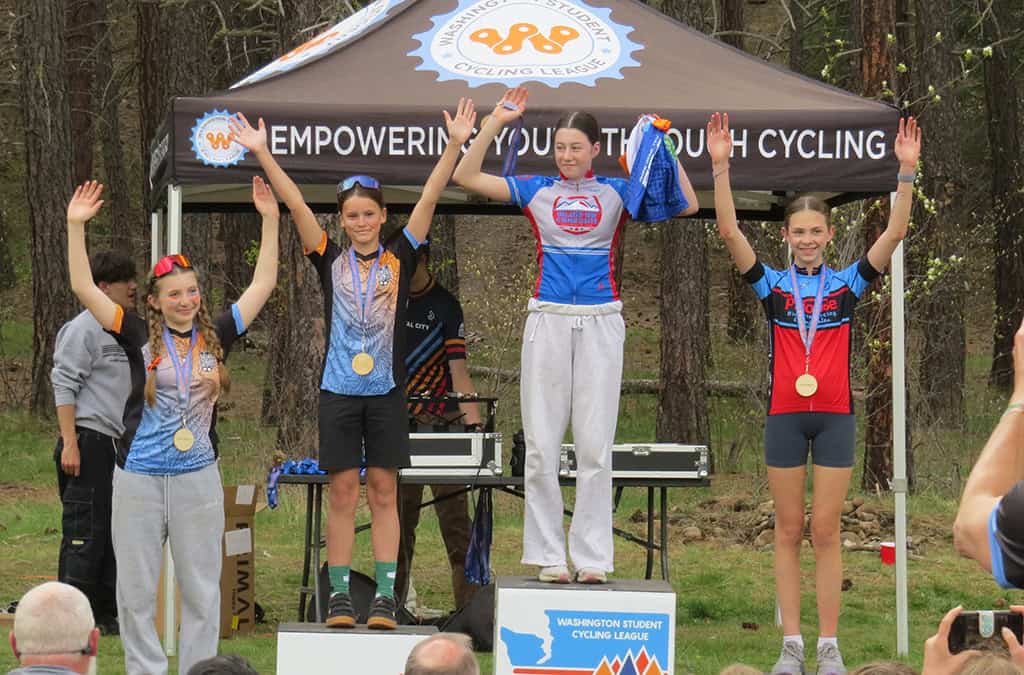 Four cyclists on the podium with medals holding hands in the air
