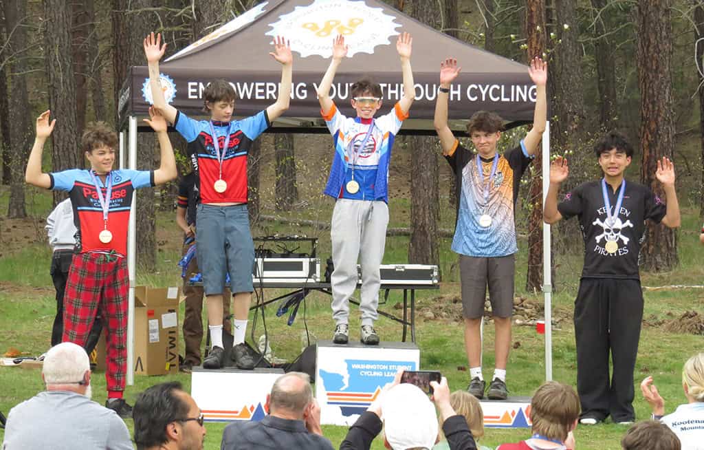 Five cyclists on podium with medals, holding hands in the air