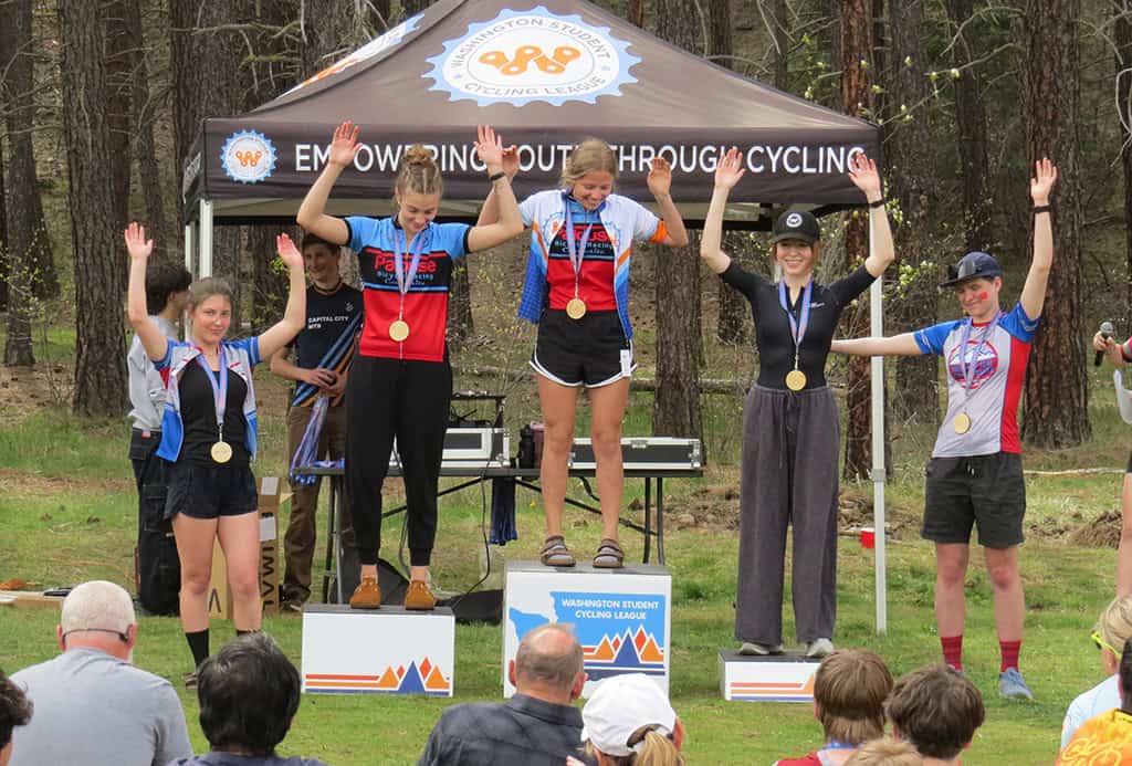 Five cyclists on podium with medals, holding hands in the air